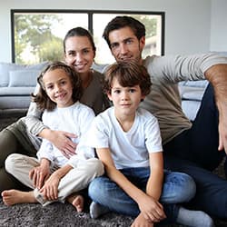 A mother, father and their two young children sitting together on the floor of their living room.